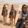 Happy tourists riding camels in the Dubai desert near a traditional camp during a group safari tour.