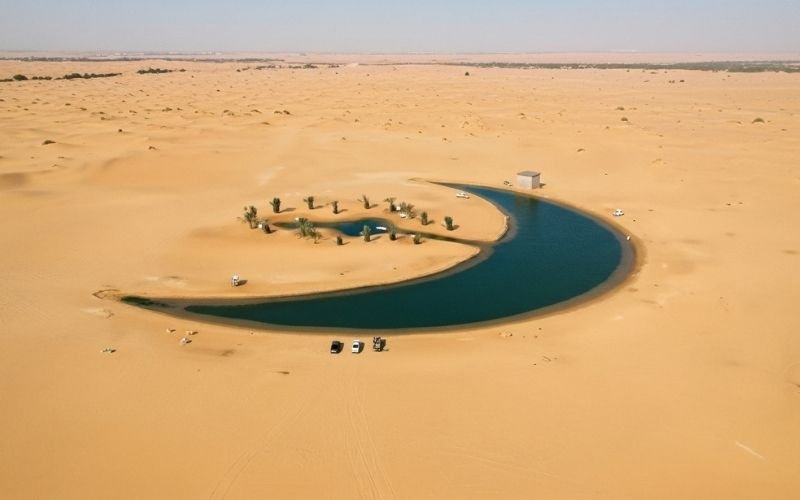 Aerial view of Moon Lake, a crescent shaped lake surrounded by golden sand dunes in Dubai’s Al Qudra Desert, with a few cars and palm trees nearby.