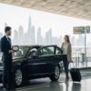 Luxury airport transfer at Dubai airport with black car, chauffeur holding name board, happy traveler with luggage, private airport transfer Dubai and DXB airport transfer service scene.